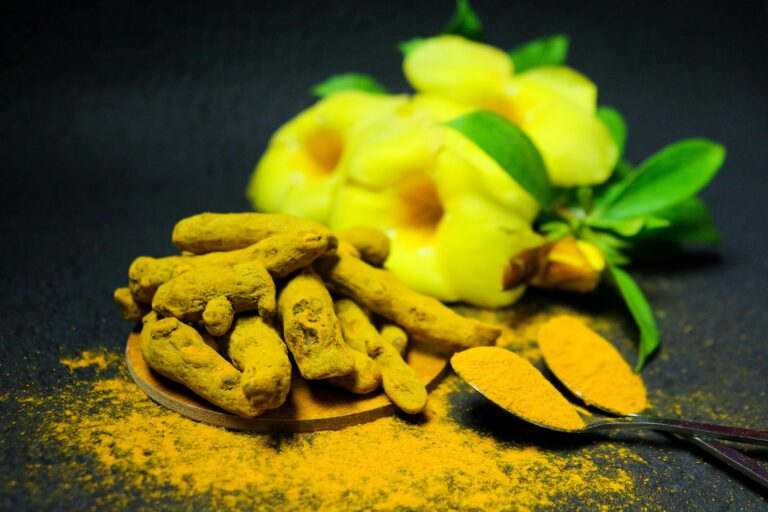 Close-up of turmeric roots and powder with green leaves and yellow flowers on black background.
